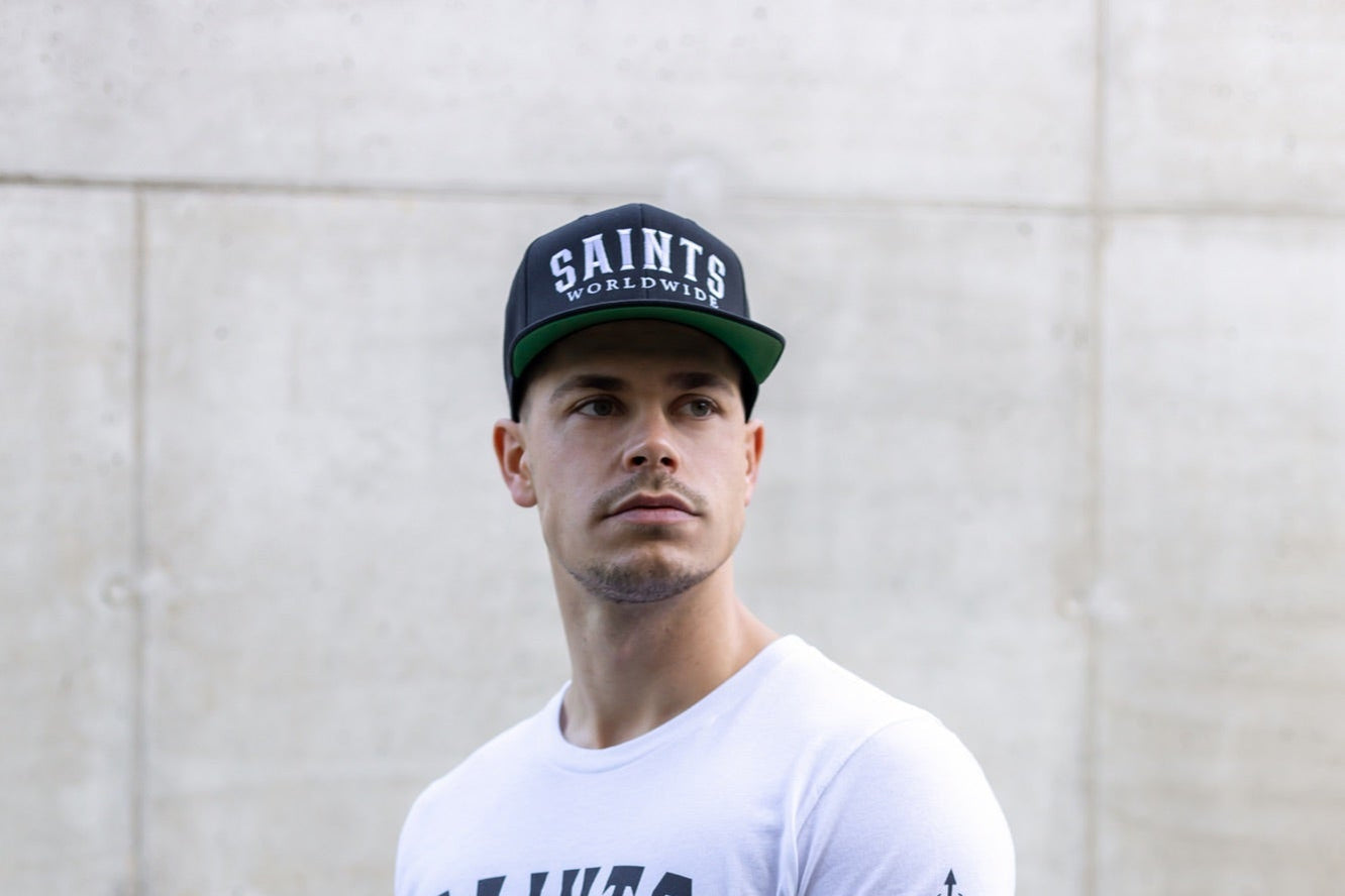 Man wearing a white SAINTS WORLDWIDE tee and black snapback against a concrete wall