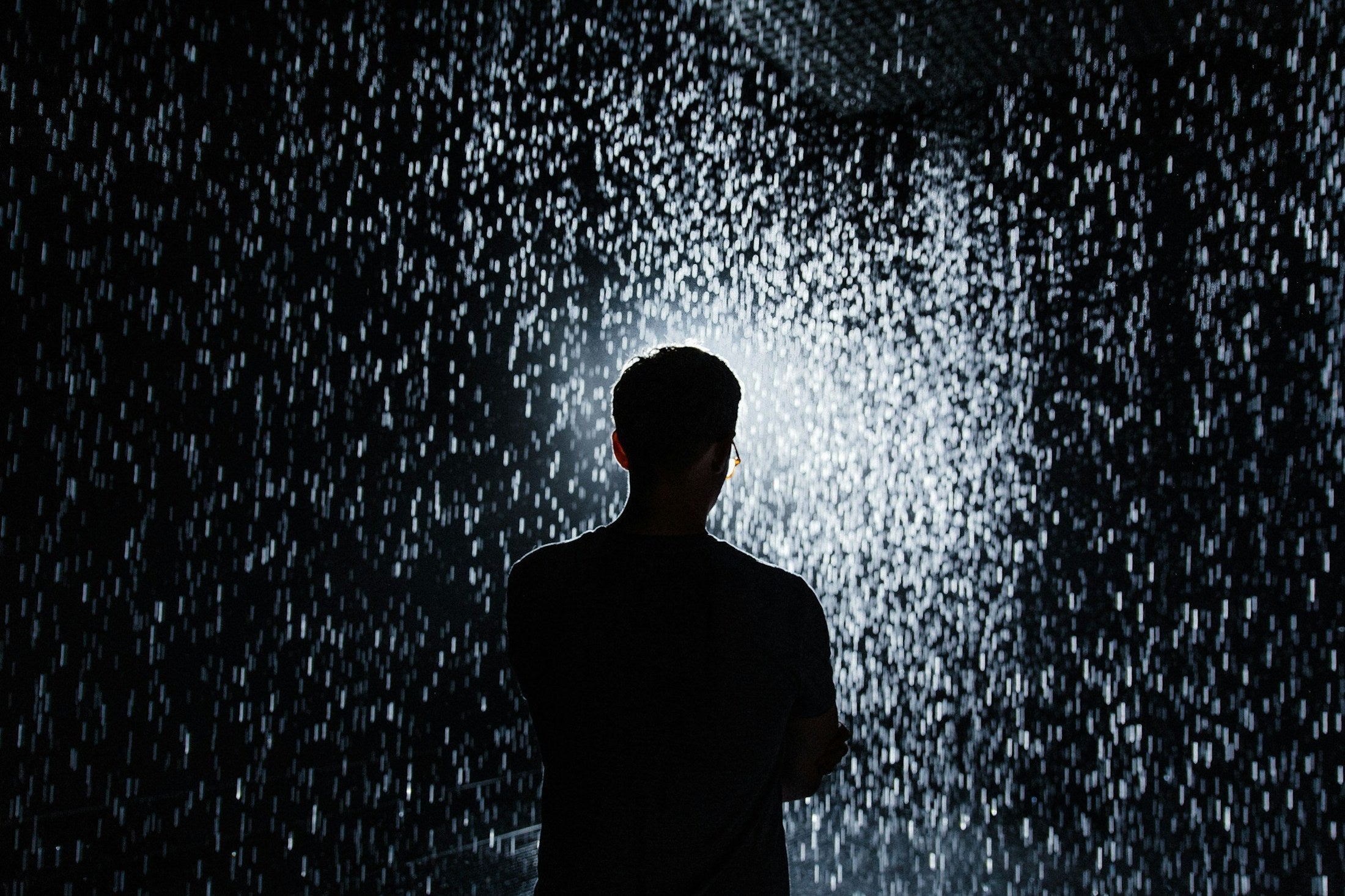A man standing still under falling rain at night, arms folded, staying under it