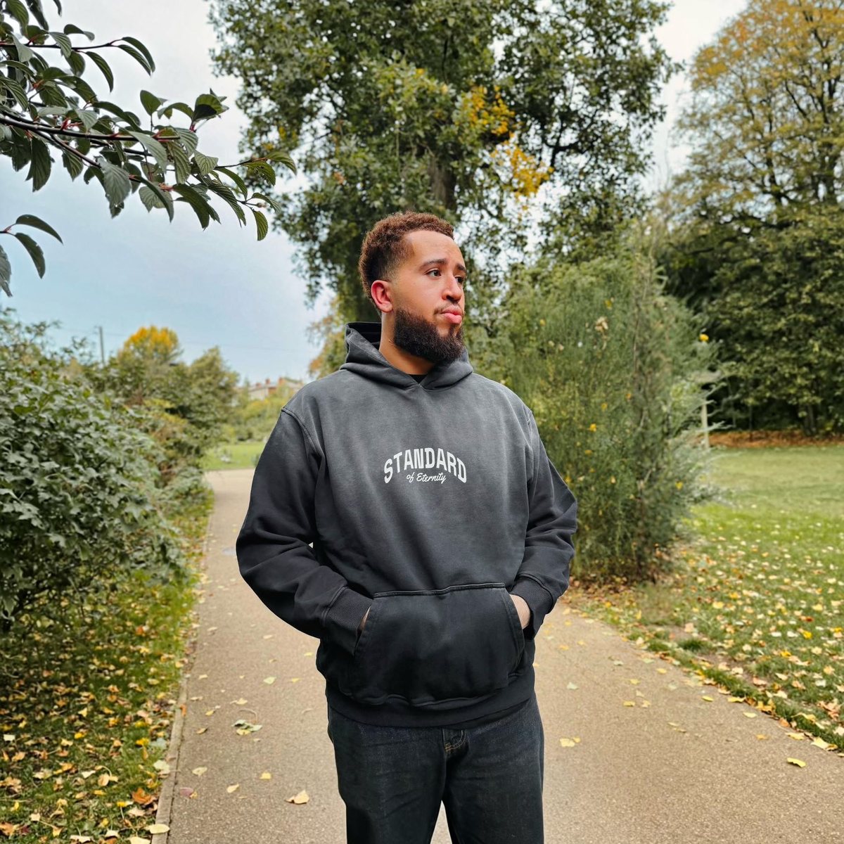 STANDARD Hoodie in washed black worn with jeans on a park path, full-length front view showing the oversized fit