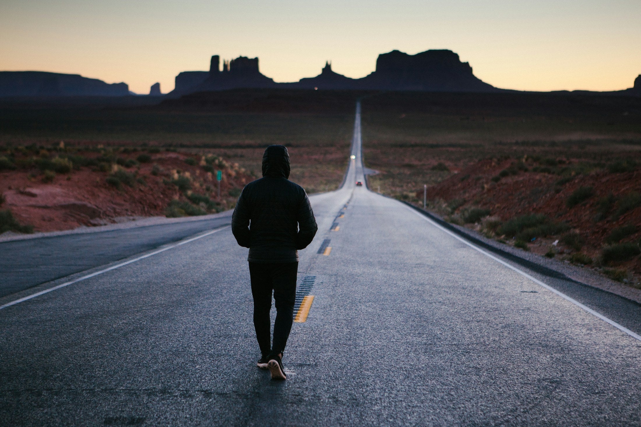 Person walking the centre line of a long open road at dusk toward the horizon